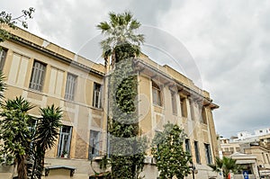 Large Neoclassic Building Facade in Downtown Helarclion, with a High Palm Tree on a Cloudy Day. Typical Street and Road with Cars