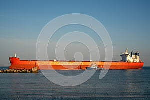 Large ocean-going cargo ship in orange. The dry cargo ship enters the port using a tug.