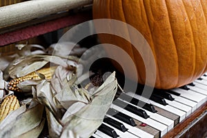 A large orange pumpkin stands on the white and black keys of an old piano next to dry ears of yellow corn.