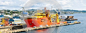 A large orange and yellow colored Offshore Construction Vessel OCV is in a dry dock of a shipyard and is being repaired