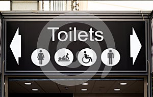 Large overhead black and white toilets sign with male, female, disabled and baby changing icons.