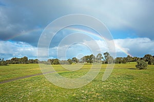 A large park with double rainbows against cloudy sky. Background texture of green grass field or meadow with Australian native