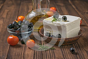 A large piece of feta cheese in a clay bowl with olives and tomatoes on a wooden table.