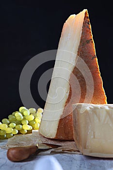 Large pieces of cheese on paper. Aged hard cheese. Grapes and a cheese knife. Black background. Vertical photo.