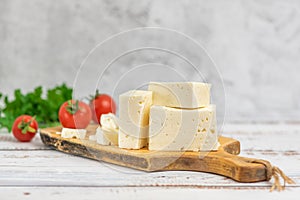 Large pieces of feta cheese on old wooden cutting board and cherry tomatoes on light background. Selective focus