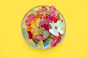 A large pile of multi-colored jelly gummy candies in a glass bowl on a yellow background