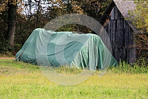 Large pile of stacked round hay bales completely covered with thick green nylon prepared for cold winter days next to wooden barn