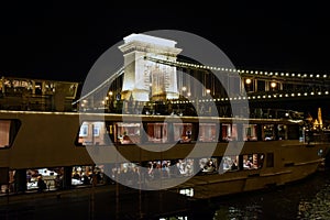 A large pleasure boat with resting people floats in front of the illuminated night bridge