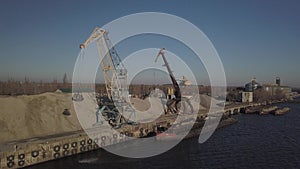 Large port cranes on the shore near the pier unload the river sand barge onto a large heap. The train for the transport of bulk ca
