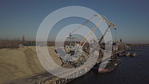 Large port cranes on the shore near the pier unload the river sand barge onto a large heap. The train for the transport of bulk ca