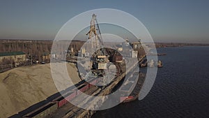 Large port cranes on the shore near the pier unload the river sand barge onto a large heap. The train for the transport of bulk ca