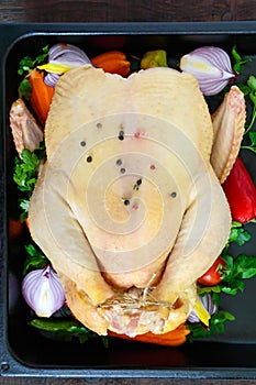 A large raw chicken with vegetables, greens in black metal form on a wooden background.