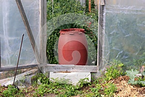 A large red barrel of water stands in the door of the wooden greenhouse