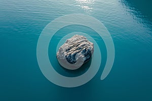 A large rock stands alone in the center of a vast body of water, surrounded by ripples and reflections, Single isolated rock in a