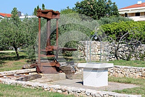 Large rusted metal vintage retro olive oil press machine mounted on concrete foundation next to newly built well used as garden