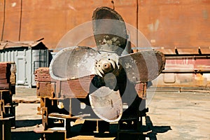 Large, rusted ship propeller displayed on wooden blocks in a shipping yard, symbolizing industrial maritime decommission