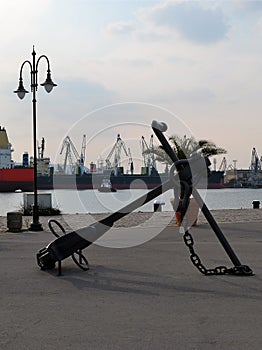 A large sea anchor adorns the walking part of the port, in the background are the water area of the port, cargo ships and cra