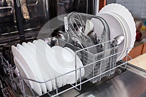 Large and small white plates, steel forks, spoons in open dishwasher