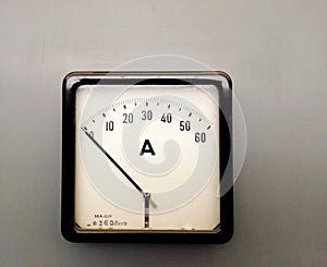 a large square industrial ammeter with an analogue dial with numbers with standard electrical symbols on a white dial on a grey