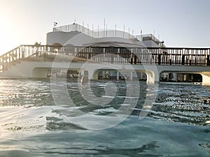 A large stone building with a pedestrian bridge with arches above the pool near the water, a pool against a blue sky and a big sun