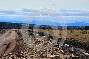 Large stones are piled on a country road that passes by a small village. There are two trucks on the road. Beautiful sky with clou