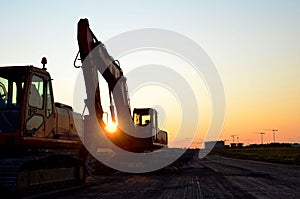 Large tracked excavator with with hydraulic hammer breaks asphalt at a construction site against on the background sunset.