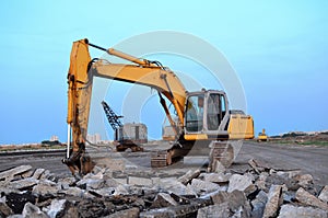 Large tracked excavator with with hydraulic hammer breaks asphalt at a construction site against on the background sunset