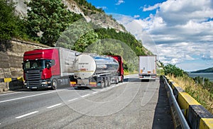 Large trucks carrying different products, on a beautiful mountain road, on a summer day. Near the road is a beautiful river, the