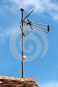 Large TV antenna mounted on top of rusted metal pole on edge of family house roof covered with dilapidated old roof tiles