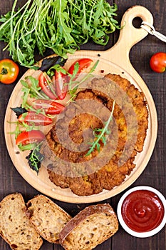 A large Viennese schnitzel and tomato salad on a cutting board on a dark wooden background.