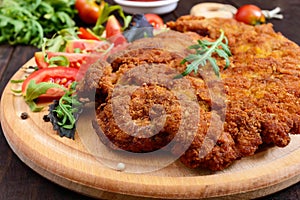 A large Viennese schnitzel and tomato salad on a cutting board on a dark wooden background.