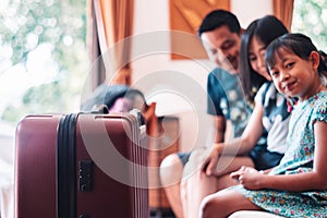 Large wheeled suitcase standing on the floor in the hotel room with happy asian family sitting on the bed in background