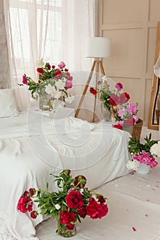 Large white bed in a bright room decorated with vases of bright peonies. Bedroom interior decorated with spring pink