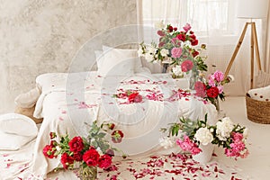 Large white bed in a bright room decorated with vases of bright peonies. Bedroom interior decorated with spring pink