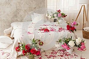 Large white bed in a bright room decorated with vases of bright peonies. Bedroom interior decorated with spring pink