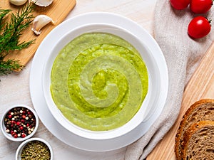 Large white bowl with vegetable green cream soup of broccoli, zucchini, green peas on white background, top view