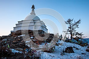 A large white Buddhist stupa against a blue sky and many colorful ribbons on bushes on the sacred island of Ogoy on Lake Baikal.