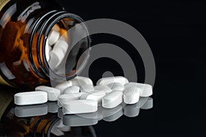 Large white capsule-shaped tablets in a dark glass bottle on a black mirror background.