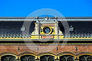 large white clock in decorative exterior yellow brick wall. black roman numerals. gray mansard zink roof