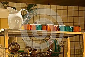 A large white jug and a row of colored mugs and cups on a shelf in the kitchen