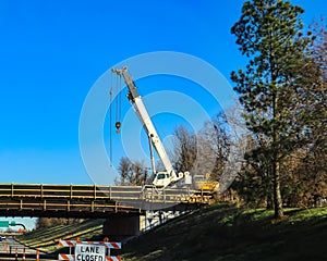 Large white mobile crane working on bridge above a highway with lane closed by tall pine tree