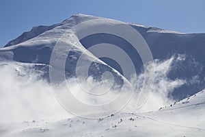 A large white mountain with a dark couloir is covered with snow among the foggy clouds before winter blizzard