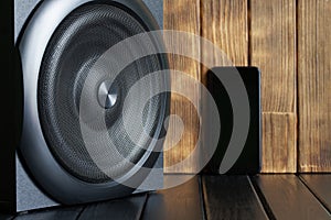 Large wireless audio speaker with a metal grill next to a smartphone against a background of natural boards. Stream music, sound