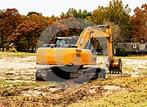 Large yellow backhoe with tracks sitting in vacant lot in leafy residential area with nice swingset and house in background