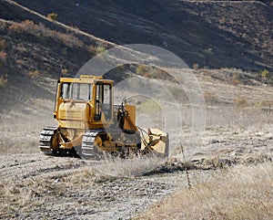 A large yellow bulldozer flattens a mountain dirt road in clouds of dust on a Sunny autumn day