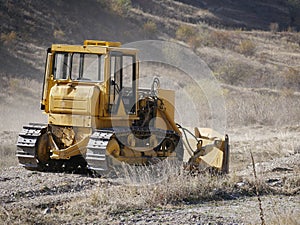 A large yellow bulldozer flattens a mountain dirt road in clouds of dust on a Sunny autumn day