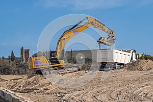Large yellow excavator depositing sand in a truck bed at a large construction site