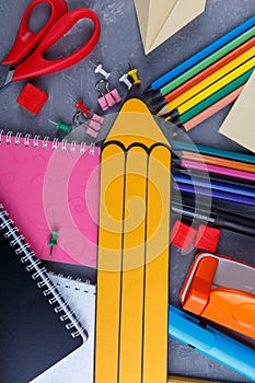 A large yellow paper pencil, next to a variety pencils, notebooks, clamps and crayons and other office supplies.