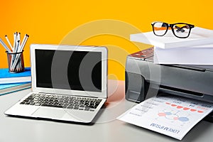 Laser printer and laptop on desk against yellow background