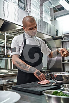 Last details. Serious bald chef in apron and with beautiful tattoos on his arms adding famous sauce to the dish while